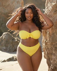 Plus size model wearing Lemon Drop yellow bandeau bikini posing on the beach near rocks in sunlight