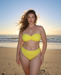 Plus size model wearing Lemon Drop yellow bandeau bikini standing on the beach at sunset with ocean waves behind