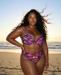 Confident plus size model in Purple Blaze Golden Hour bikini set featuring vibrant purple and orange flame print on a beach at sunset