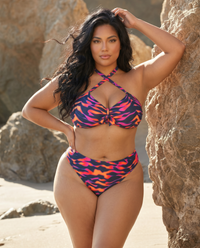 Plus size model wearing Purple Blaze flame print bikini posing on the beach near rocks with ocean backdrop