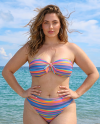 Plus size model wearing Sweet Stripes rainbow striped bandeau bikini standing in front of the ocean on the beach