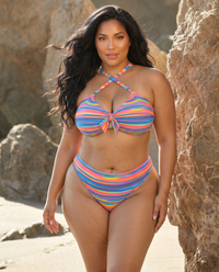 Plus size model wearing Sweet Stripes rainbow striped bikini with cross strap top posing on the beach near rocks