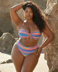 Plus size model wearing Sweet Stripes rainbow striped bikini set posing against rocks on the beach